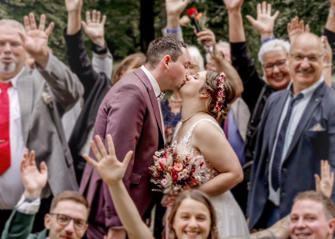 Braut und Bräutigam küssen sich umgeben von feiernden Gästen mit erhobenen Händen.
