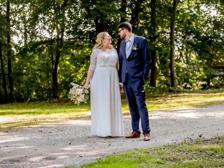 Braut und Bräutigam stehen auf einem Weg, umgeben von Bäumen, und blicken sich an.
