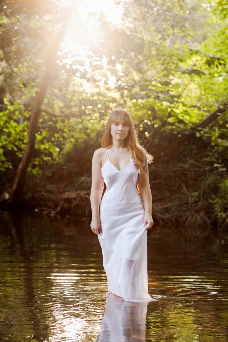 Frau in einem weißen Kleid steht im Wasser, umgeben von Bäumen und Sonnenstrahlen.