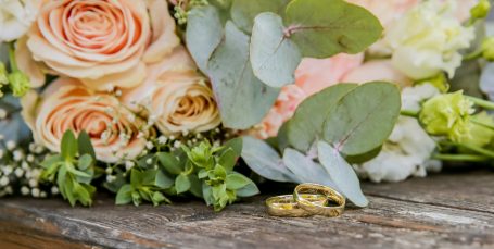 Brautstrauß mit Rosen und Eukalyptus, zwei goldene Eheringe liegen auf Holzoberfläche.