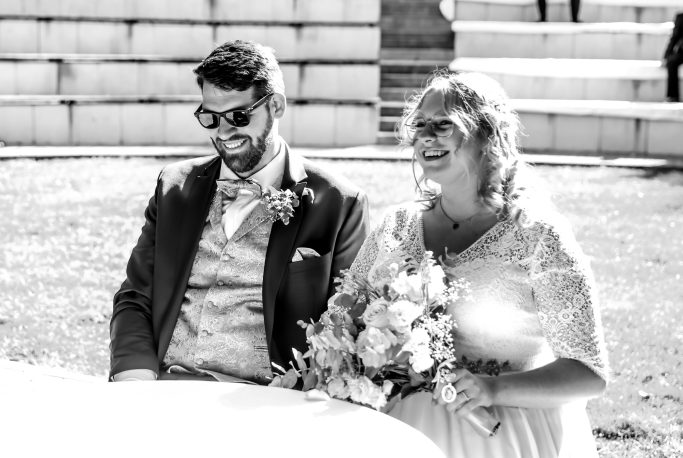 Ein Brautpaar sitzt lächelnd im Freien, umgeben von Blumen und Sonnenschein.