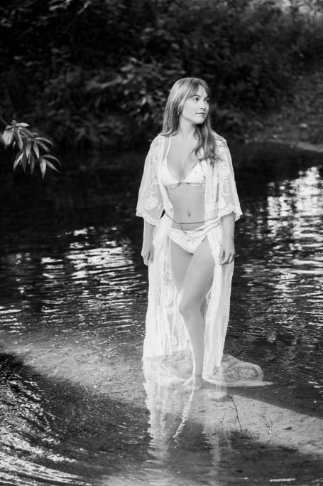 Frau in einem weißen Gewand steht im seichten Wasser umgeben von grüner Natur.