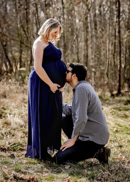 Schwangere Frau in einem langen blauen Kleid, Mann küsst ihren Bauch im Wald.