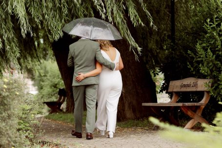 Ein Paar in eleganter Kleidung, unter einem Regenschirm, umarmt sich unter einem Baum.
