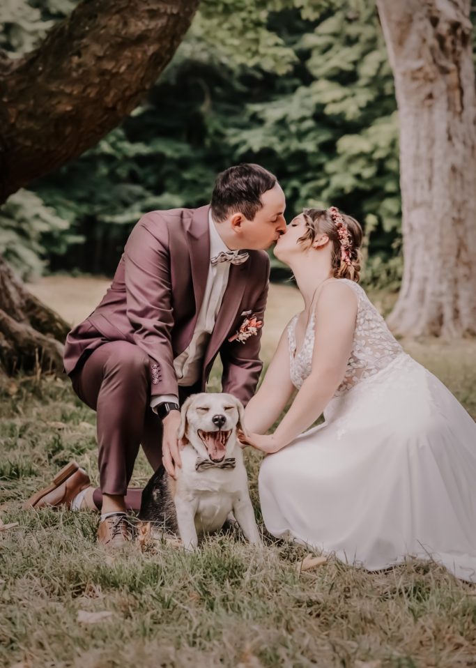 Brautpaar küsst sich im Freien, während sie neben ihrem Hund sitzen.