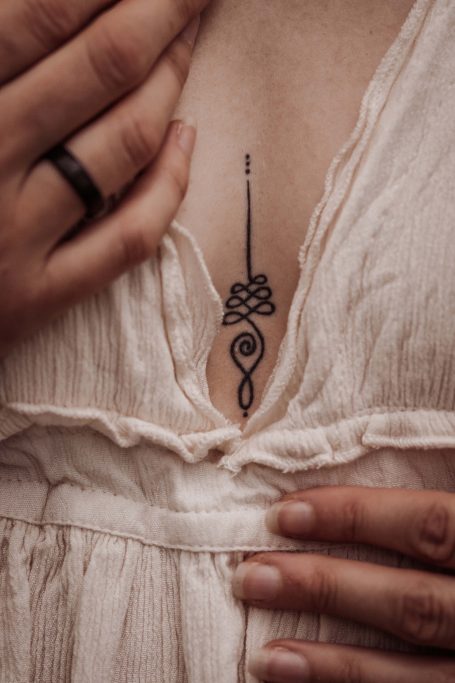 Schwarzes Tattoo in Form eines Knotens und einer Spirale zwischen den Brustlinien.