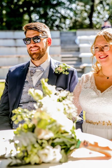 Hochzeitspaar im Freien, lächelnd mit Blumenstrauß, Braut in weißem Kleid, Bräutigam in Anzug.
