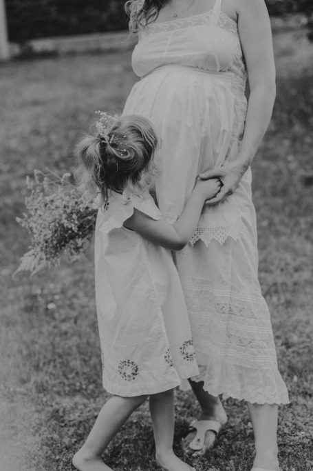 Schwangere Frau umarmt ein Mädchen, das ihr Bein hält und Blumen hält.