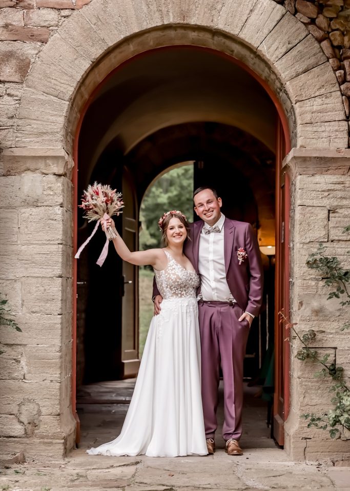 Braut und Bräutigam stehen in einem Torbogen und lächeln, Braut hält einen Brautstrauß.