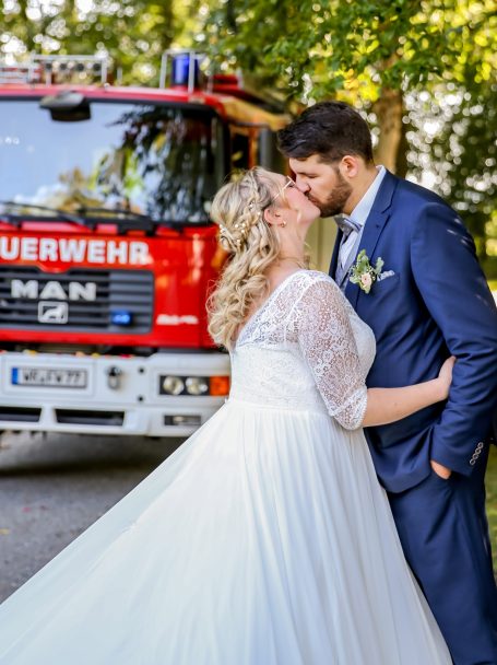 Brautpaar küsst sich vor einem roten Feuerwehrfahrzeug in der Natur.