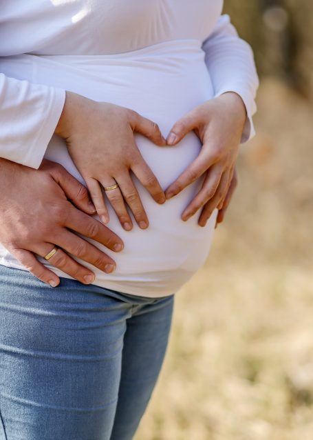 Hände bilden ein Herz über einem schwangeren Bauch im Freien.