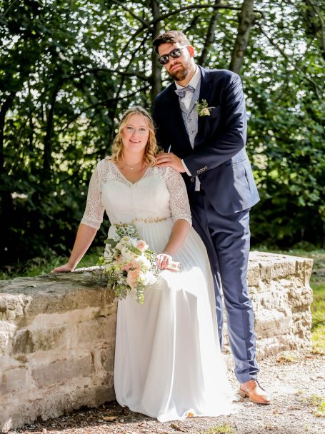 Brautpaar posiert vor einer grünen Kulisse, Braut in weißem Kleid, Bräutigam in Anzug.