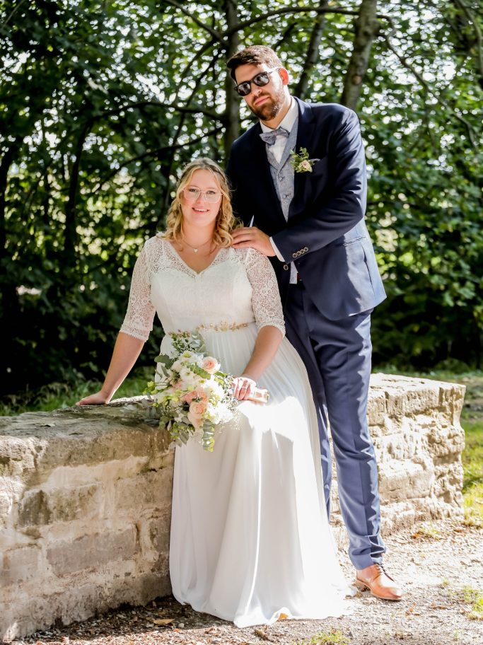 Brautpaar posiert vor einer grünen Kulisse, Braut in weißem Kleid, Bräutigam in Anzug.