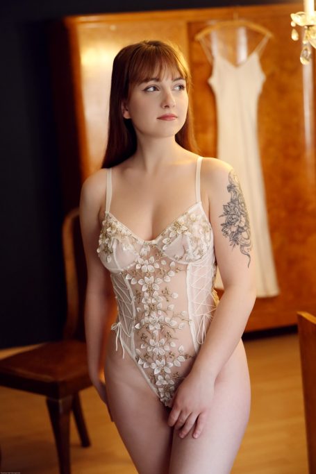 Junge Frau in einem Spitzen-Bodysuit vor einem Kleiderschrank und einem Kleid.